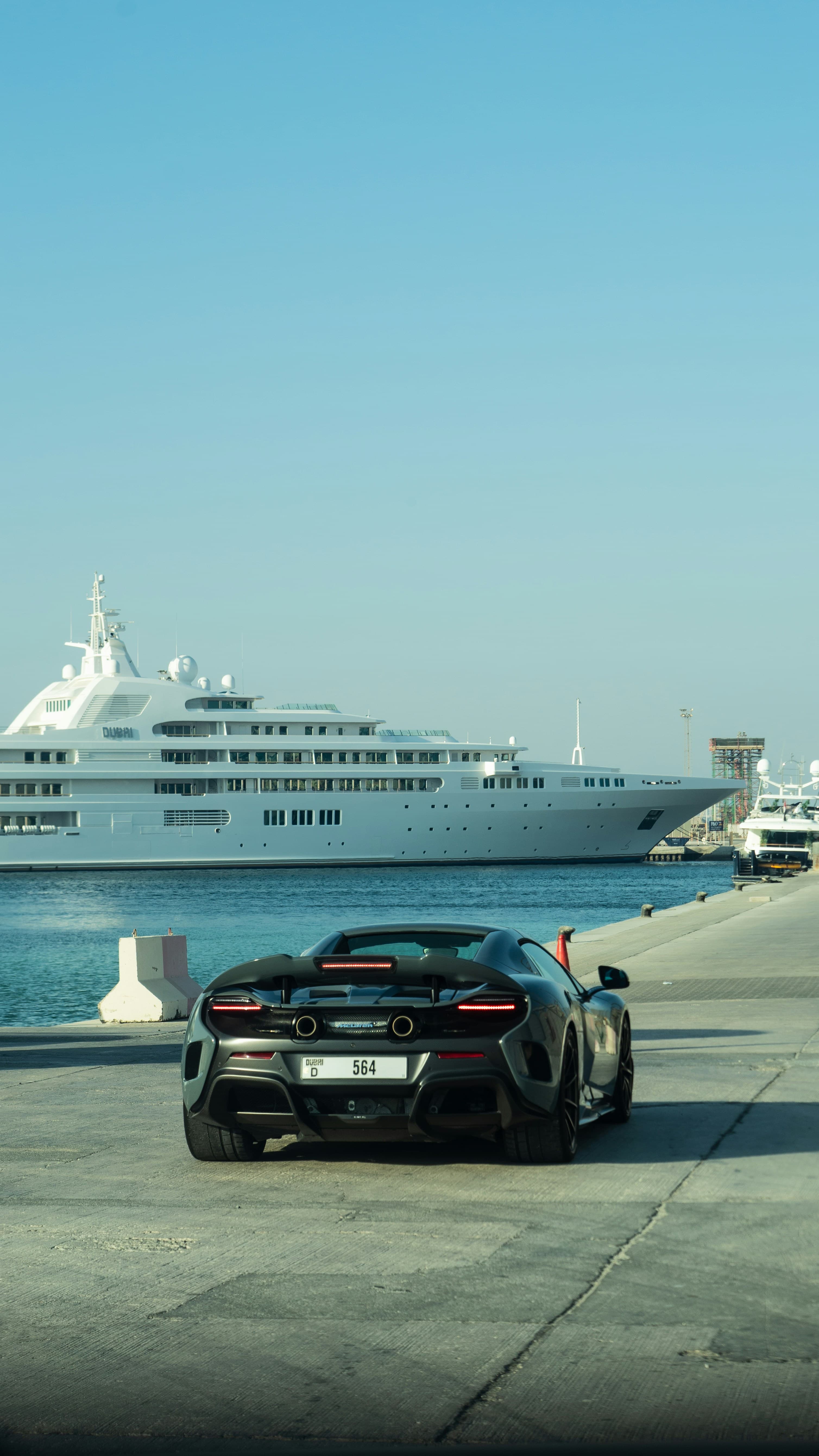 Rear view of a gray McLaren parked on a pier facing a massive white yacht.