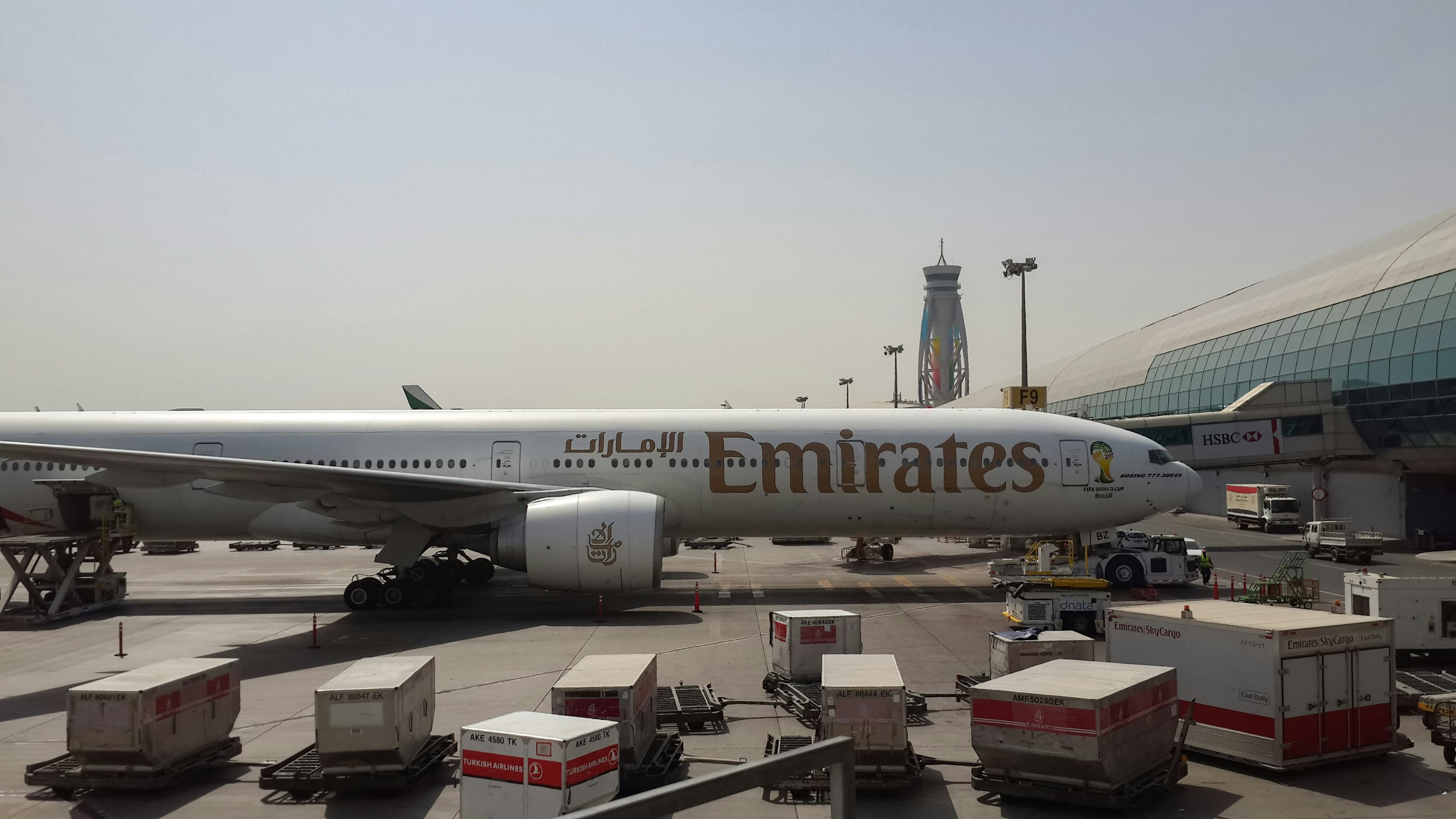 Emirates Boeing 777-300ER parked at an airport gate with cargo containers in the foreground.