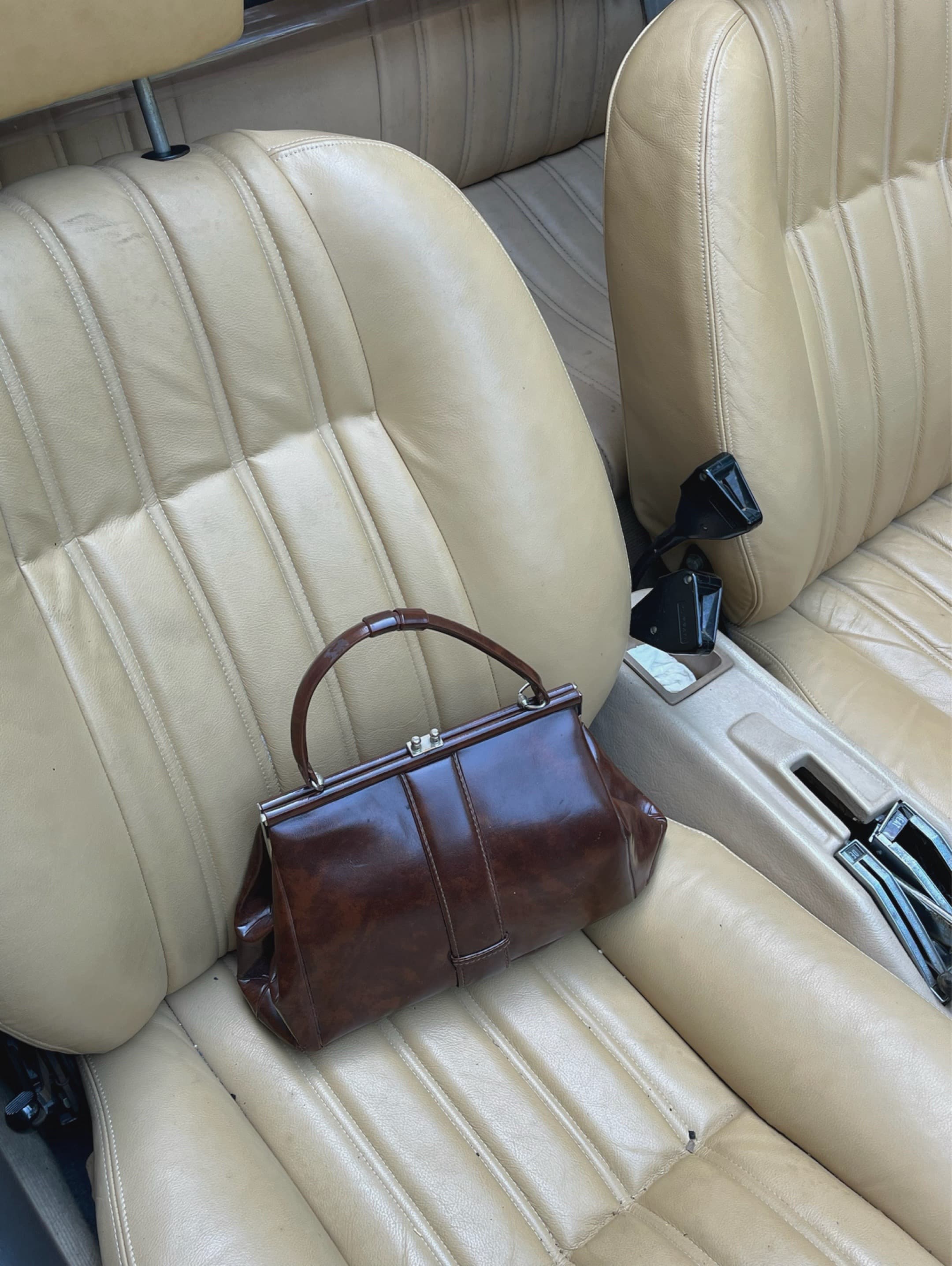 Glossy brown vintage handbag sitting on the tan leather seat of a classic car.