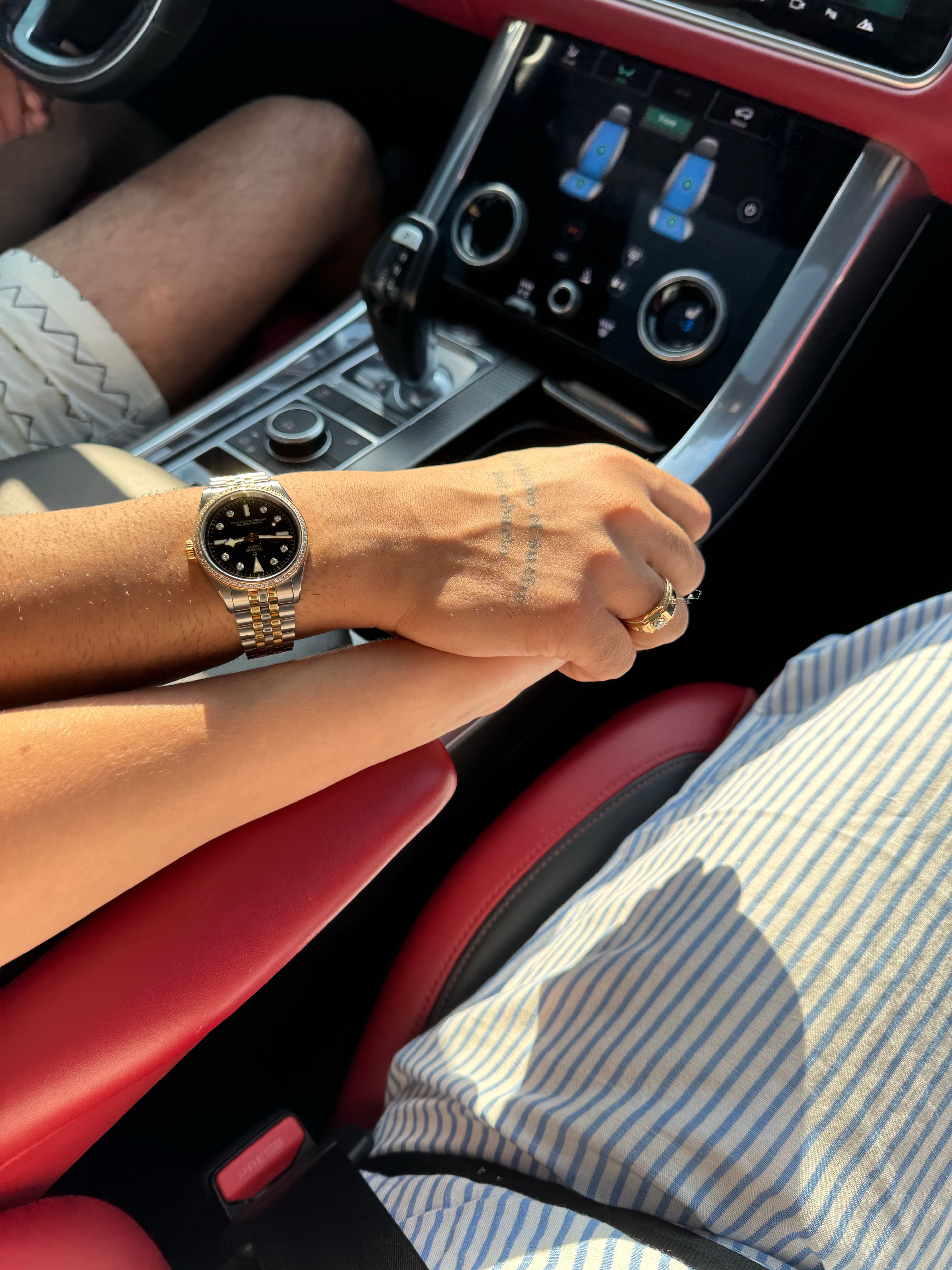 Two people holding hands in a luxury car with red leather interior and gold watch.