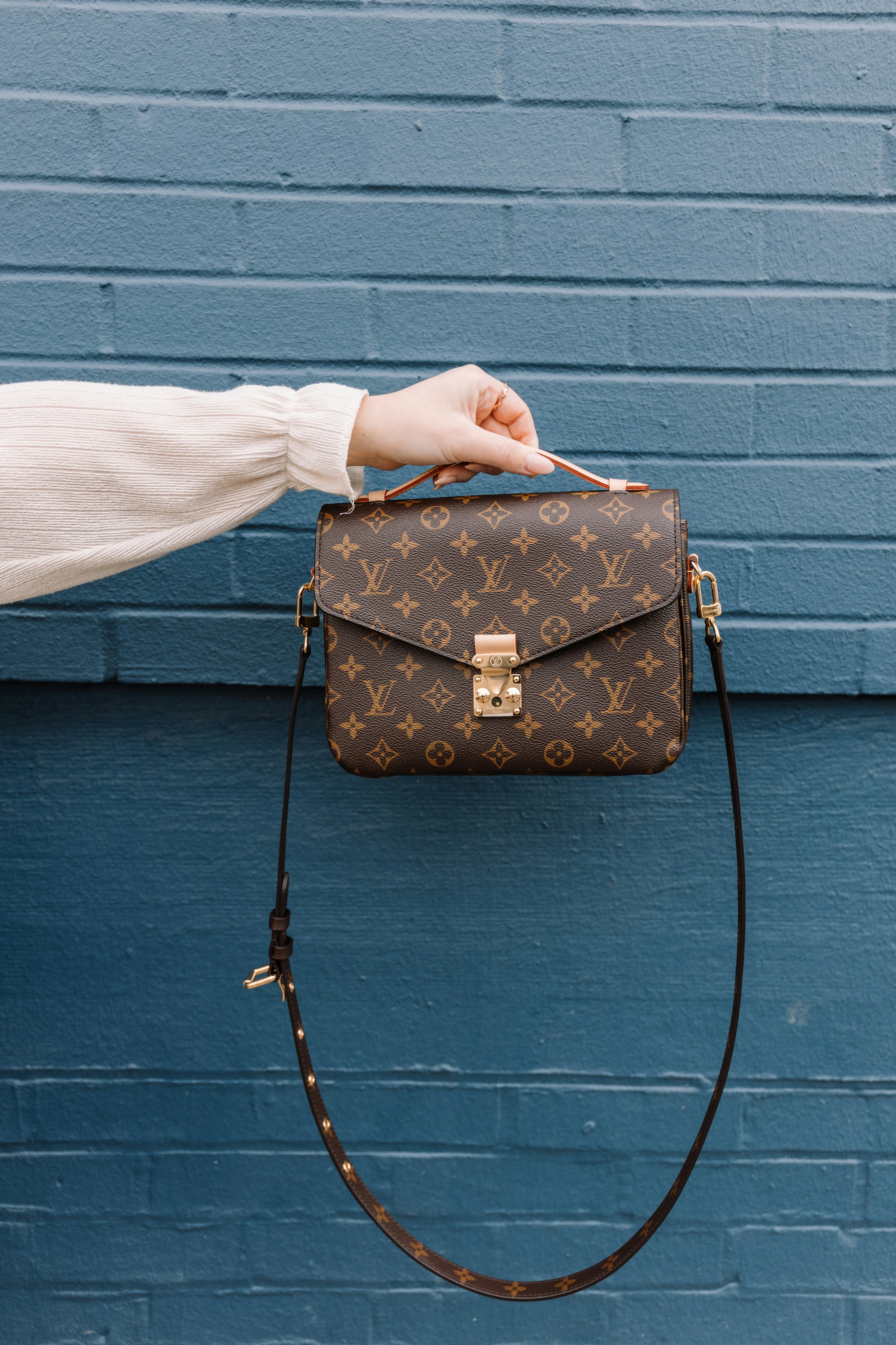 Hand holding a Louis Vuitton monogram handbag in front of a blue brick wall.