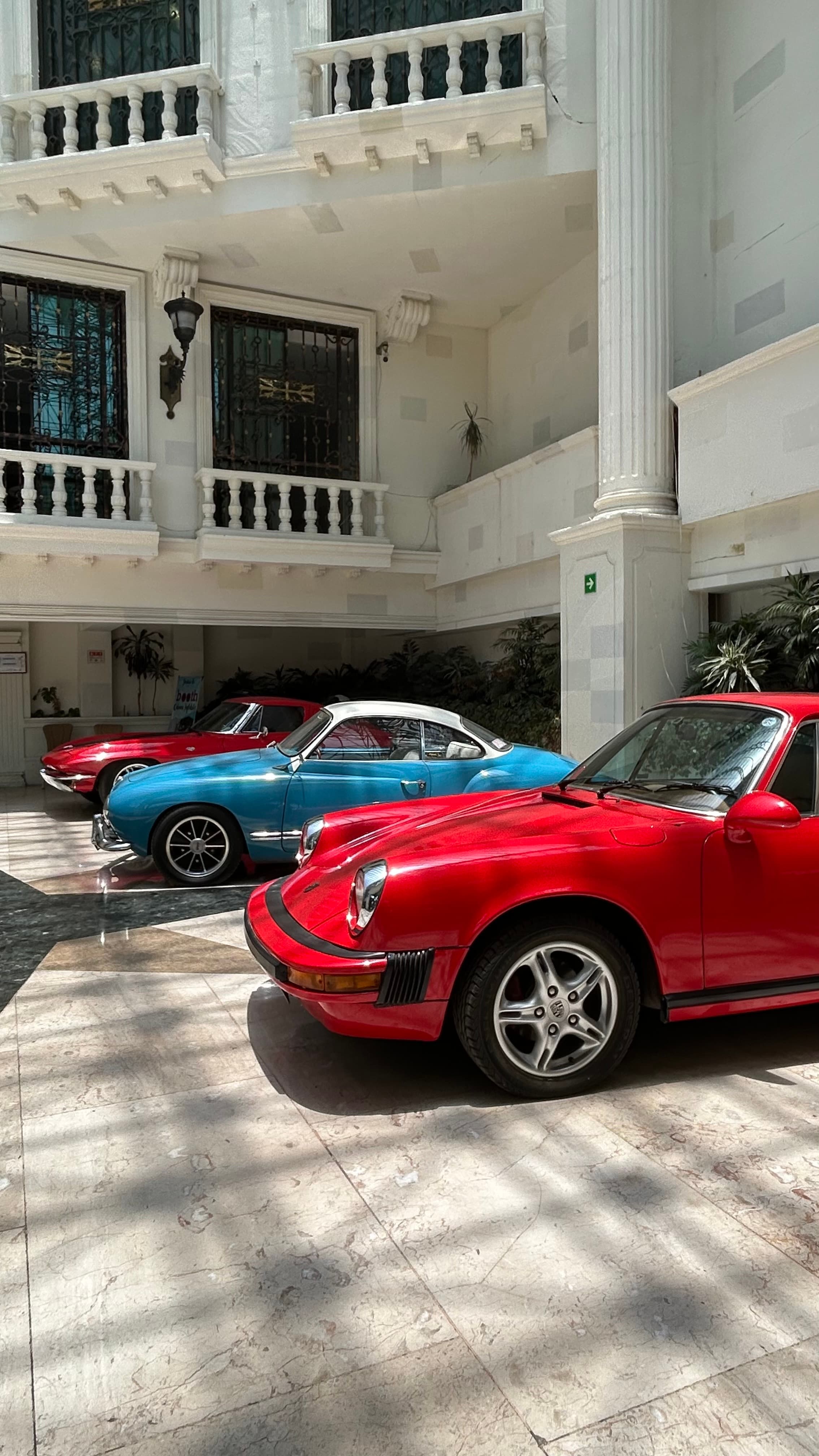 Red Porsche, blue Karmann Ghia, and red Corvette parked in an ornate white courtyard.