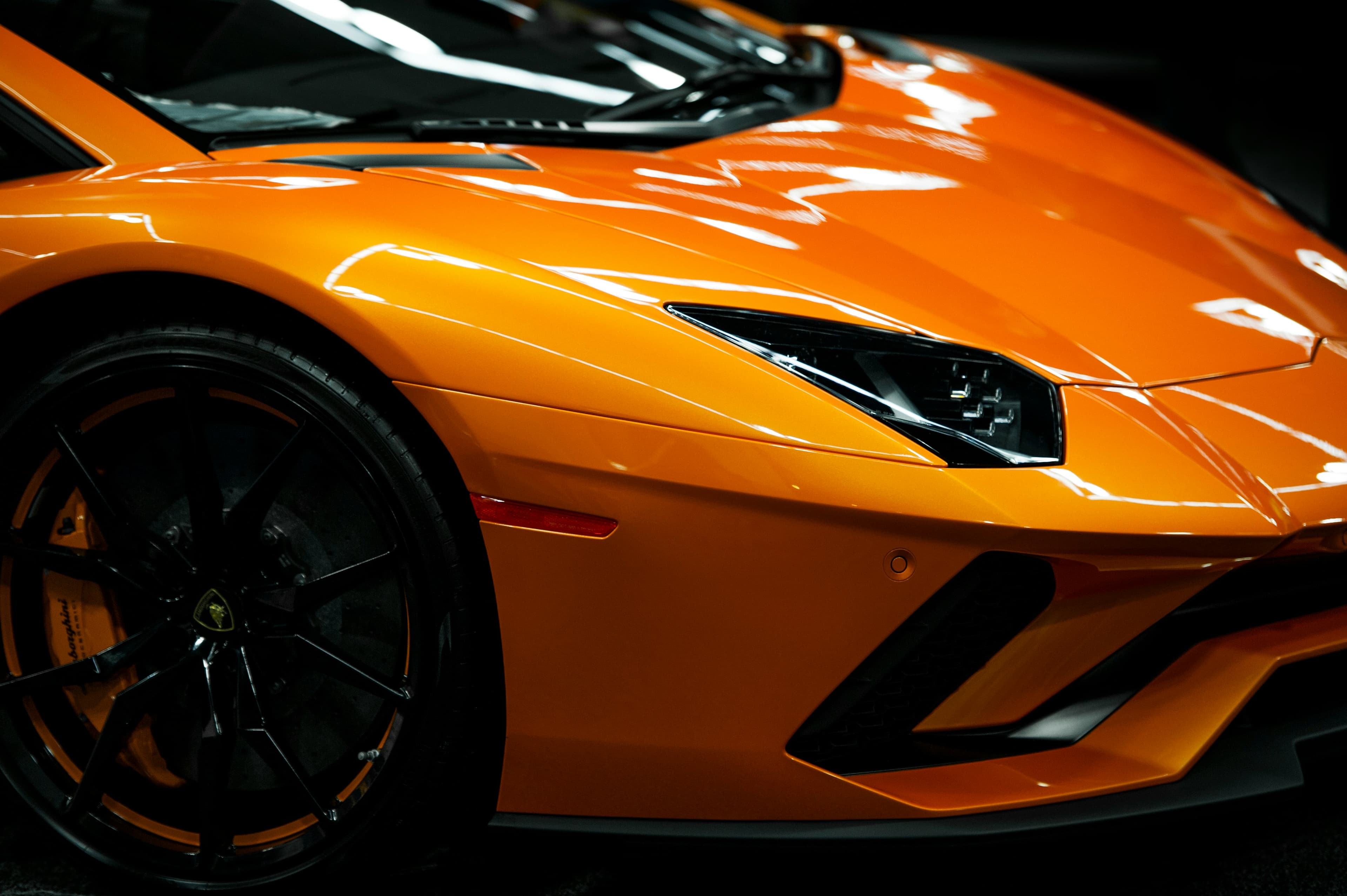 Sleek orange Lamborghini supercar with sharp body lines, angular headlights, and black multi-spoke wheels.